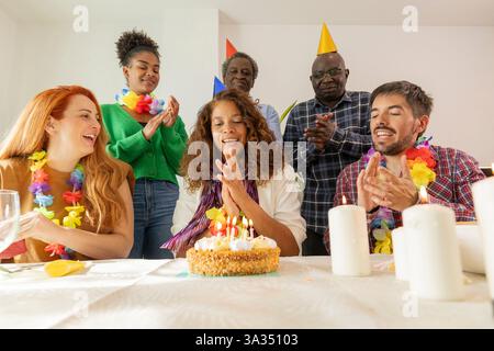 Una vivace festa di compleanno con un gruppo riunito intorno a una torta. Cappelli da festa, leis colorati e volti sorridenti creano un'atmosfera gioiosa. Foto Stock