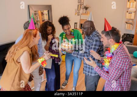 Un gruppo di amici che festeggiano un compleanno al chiuso con sorrisi e vivaci decorazioni per feste. Si riuniscono intorno a una torta, creando un'atmosfera gioiosa. Foto Stock