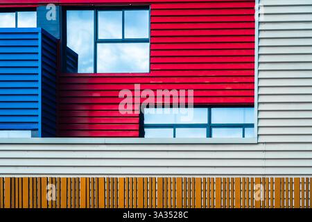 Questa immagine cattura la vibrante e moderna facciata urbana di un edificio, caratterizzato da un audace mix di pannelli rossi, blu e bianchi interlacciati con un chiaro windo Foto Stock