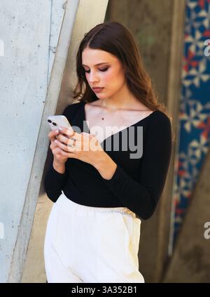 Una donna con la parte superiore nera e i pantaloni bianchi si appoggiano a una parete di cemento mentre utilizza uno smartphone. Sembra concentrata e premurosa, con un colore Foto Stock