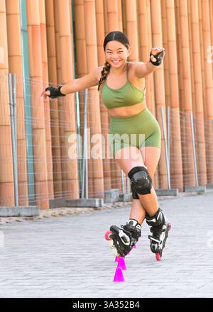 Una giovane donna cinese in verde indossa abilmente rollerblade attraverso una linea di coni. Indossa gli indumenti protettivi e sorride mentre si bilancia Foto Stock