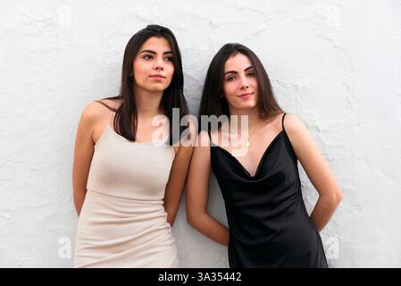 Le giovani sorelle in abiti alla moda, una in nero e l'altra in bianco, si stagliano contro un muro bianco testurizzato, trasudando eleganza e amicizia. Foto Stock