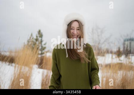 Una donna gioiosa con un maglione verde e un caldo cappello bianco invernale sorride in un parco innevato. Erba alta e alberi sullo sfondo offrono un inverno sereno Foto Stock
