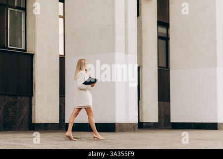 Un professionista in un abbigliamento formale cammina con fiducia tenendo in mano una cartella, adagiato su un moderno edificio per uffici, incarnando l'eleganza aziendale Foto Stock