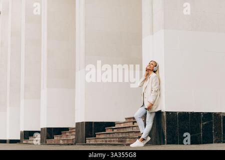 Una donna d'affari si appoggia a una colonna di un edificio, ascoltando musica con le cuffie. Vestita in modo informale con una giacca e un paio di jeans, ha un look rilassato Foto Stock
