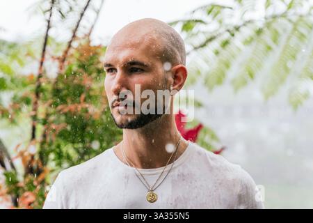 Un uomo sta sotto la pioggia, nel profondo del pensiero, circondato da una vegetazione lussureggiante in una foresta. Le gocce cadono intorno a lui, creando un'atmosfera contemplativa e serena Foto Stock