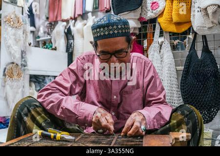 Un artigiano balinese che indossa occhiali si concentra sulla lavorazione di prodotti fatti a mano in un vivace mercato locale. Borse a uncinetto colorate adornano lo sfondo e riflettono Foto Stock