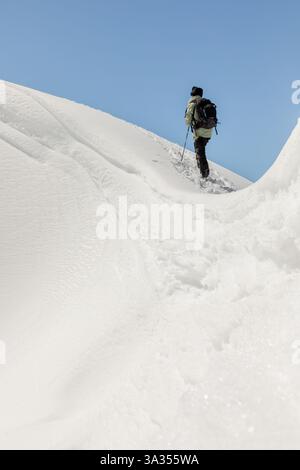 Vista posteriore di un arrampicatore irriconoscibile che sale su un pendio di montagna ripido e innevato, utilizzando pali per sostenerlo sotto un cielo azzurro. Foto Stock