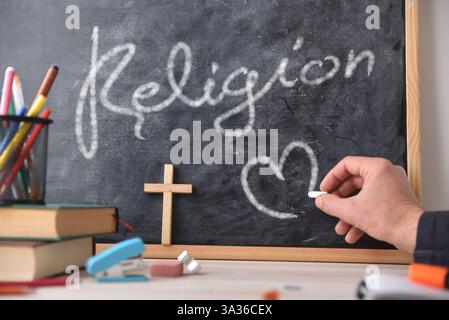 Disegnare a mano un cuore con il gesso su una lavagna con la parola religione scritta sopra. Una scrivania di legno con libri e materiale scolastico. Vista frontale. Foto Stock