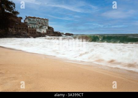 Una lunga esposizione cattura la potenza dell'oceano mentre un'onda si schianta sulla spiaggia sabbiosa di fronte a un edificio Foto Stock