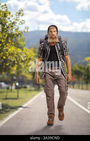 Ritratto a figura intera di un giovane escursionista barbuto con uno zaino che cammina in un parco cittadino Foto Stock