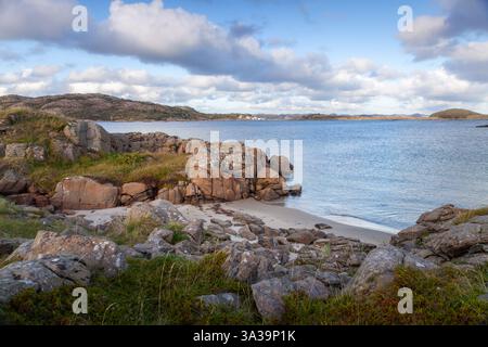 Un tranquillo paesaggio costiero presenta una costa rocciosa con sabbia dorata, scintillanti acque calme e soffici nuvole che si spostano attraverso un cielo luminoso a Farsund Foto Stock