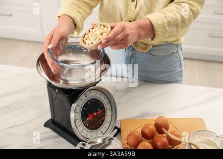 Donna che aggiunge arachidi nel recipiente su una bilancia meccanica da cucina al tavolo di marmo bianco all'interno, primo piano Foto Stock