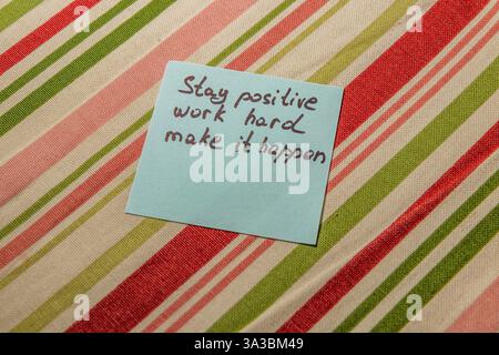 Una nota scritta a mano che mostra le frasi rimane positiva, lavora sodo e fa in modo che accada poggia su un tessuto a righe. Il messaggio ispira la produttività e Foto Stock