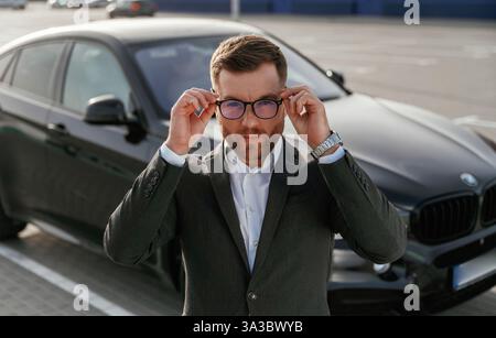 Con gli occhiali, vista frontale. L'uomo d'affari in tuta e' vicino alla sua auto nera all'aperto. Foto Stock