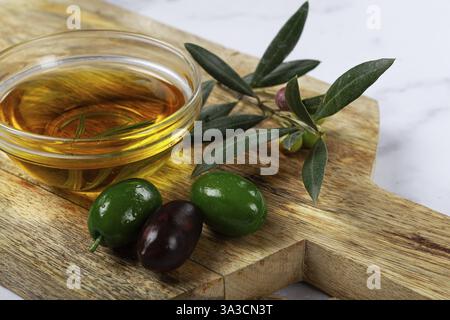 Una ciotola di vetro piena di ricco olio d'oliva si trova su un tagliere di legno accanto a olive verdi e nere fresche, completate da foglie d'oliva, creando Foto Stock