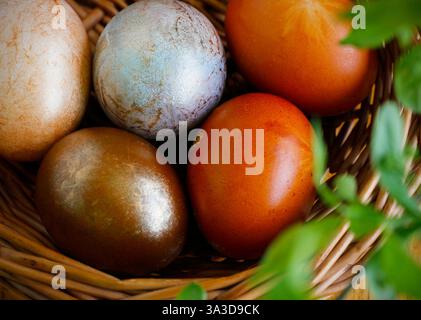 Cinque uova di Pasqua dipinte in diverse sfumature di colori neutri giacenti in un cesto di vimini sotto un ramoscello primaverile con foglie verdi fresche. Foto Stock