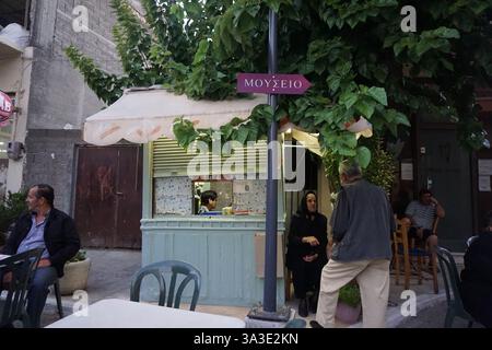 Una donna anziana siede all'interno di un chiosco e fuori un'altra sta parlando con un uomo, altri uomini sono seduti intorno - Un cartello con una freccia dice "museo" Foto Stock