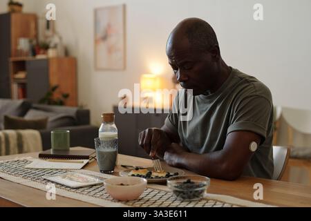Uomo afroamericano che fa una sana colazione al tavolo di legno in un soggiorno minimalista con illuminazione soffusa e arredamento accogliente. Vari piatti per la colazione e una bevanda vista sul tavolo Foto Stock