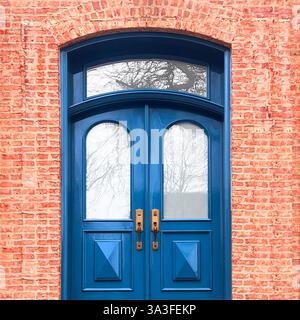 Eleganti porte doppie blu ad arco con pannelli in vetro, poste all'interno di una facciata rustica in mattoni rosa, con dettagli in ottone vintage. Foto Stock