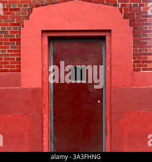 Una porta in acciaio rosso intenso con una piccola finestra quadrata si erge incorniciata da audaci murature rosse e ricchi mattoni in un vicolo di Chicago. Foto Stock