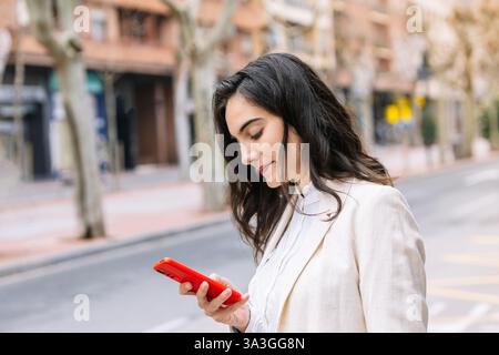 Una donna professionista in un blazer beige guarda il suo smartphone mentre cammina per una strada urbana. sembra concentrata e impegnata, usando il suo dispositivo Foto Stock