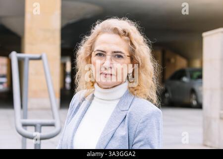 Donna matura professionale con capelli e occhiali biondi ricci, con un blazer grigio chiaro e dolcevita bianco, all'esterno di una moderna struttura da ufficio Foto Stock