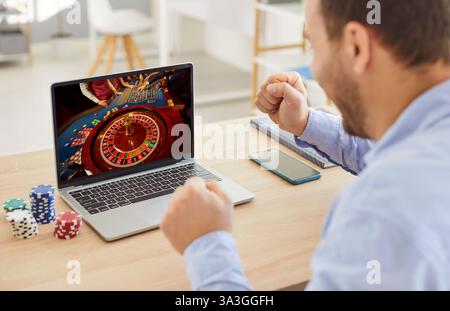 L'uomo reagisce con gioia, alzando i pugni in trionfo dopo una mano di successo mentre gioca a poker online. Foto Stock