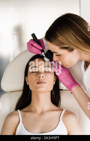 Il dermatologo esamina talpe e segni di natalità di una ragazza paziente utilizzando il dermatoscopio. Dermatoscopia di lesioni cutanee e nei con dermatoscopio per la prevenzione Foto Stock