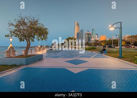 16 gennaio 2025, Abu Dhabi, Emirati Arabi Uniti: Passeggiata vuota con lampioni illuminati e grattacieli sullo sfondo al tramonto Foto Stock