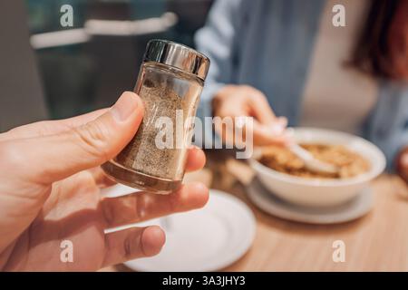 Primo piano del peperoncino in mano, pronto per condire il cibo, con persone sfocate che mangiano sullo sfondo Foto Stock