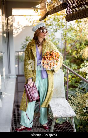 Una donna alla moda in un outfit chic si posa con un bouquet di tulipani su un elegante patio. Vestita con un cappotto verde, pantaloni a righe e tacchi rossi, trasuda fiducia ed eleganza in un ambiente naturale urbano Foto Stock