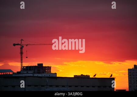 Una gru da costruzione si staglia contro un cielo rosso e arancione intenso. L'orizzonte luminoso crea un contrasto sorprendente con l'elem urbano scuro Foto Stock