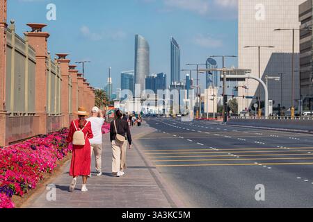 19 gennaio 2025, Abu Dhabi, Emirati Arabi Uniti: I pedoni passeggiano lungo un vivace marciapiede fiancheggiato da fiori ad Abu Dhabi, con il moderno skyline della città e i grattacieli Foto Stock