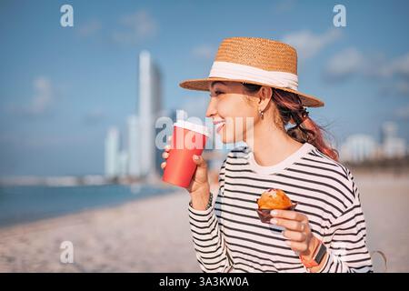 Giovane donna che fa colazione con caffè e muffin sulla spiaggia pubblica con lo skyline di Abu Dhabi sullo sfondo Foto Stock