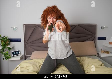 Giovane donna che canta con lo smartphone a letto e ti punta il dito contro Foto Stock