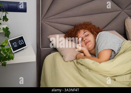 Giovane donna che dorme tranquillamente a letto, orologio digitale che mostra mezzogiorno Foto Stock