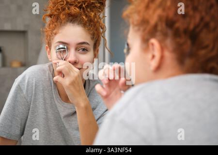Giovane donna che usa l'arricciacapelli ciglia nello specchio del bagno Foto Stock