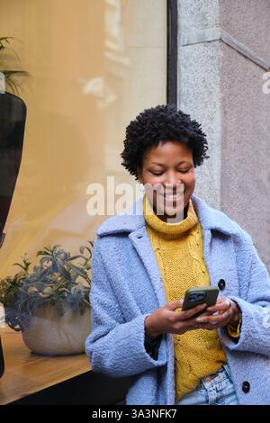 Giovane donna nera che usa il telefono e sorride mentre sta in piedi in strada Foto Stock