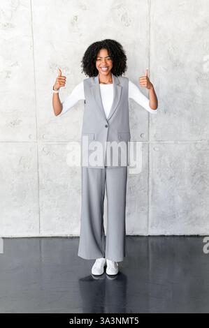 Un'allegra donna d'affari afroamericana sta con i pollici in alto, il suo sorriso vibrante e il suo gesto positivo indicano approvazione e fiducia in un ambiente professionale. Foto Stock