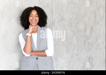 Una gioiosa donna d'affari afro-americana con un'espressione giocosa, posa con cura, riflettendo una miscela di professionalità e carisma amichevole in un ambiente aziendale Foto Stock