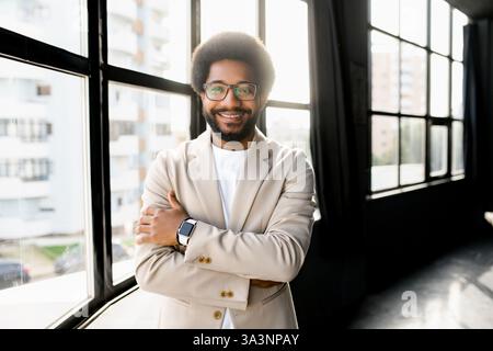 Un giovane uomo d'affari brasiliano con una moderna tuta beige si erge con le braccia incrociate, trasudando fiducia e disponibilità sullo sfondo di un ambiente di ufficio urbano. Foto Stock
