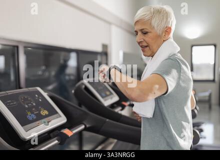 Donna anziana sorridente che corre sul tapis roulant in una palestra ben attrezzata, indossa cuffie e un asciugamano avvolto intorno al collo, guardando un orologio intelligente. Foto Stock