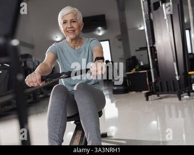 Senior femminile si sta allenando su vogatore nel moderno centro fitness, focalizzato sull'esercizio fisico per la salute Foto Stock
