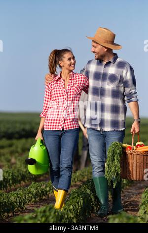 Uomo e donna contadini sorridenti mentre camminano attraverso un lussureggiante orto, portando un cesto di prodotti freschi e annaffiatoio sotto il cielo azzurro Foto Stock