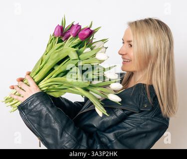 Una donna bionda allegra con una giacca in pelle nera racchiude un grande bouquet di tulipani bianchi e viola. Sorride mentre guarda i fiori contro un Foto Stock
