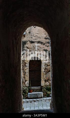 Un'antica porta in legno con arco in pietra si erge alla fine di un passaggio buio, con pareti in pietra danneggiate in un vicolo storico del villaggio. Una pentola con una pianta Foto Stock