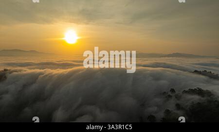 Vista aerea mozzafiato del sole che sorge su una valle di montagna coperta da un mare denso di nuvole ondulate. La luce dorata illumina la nebbia, cre Foto Stock
