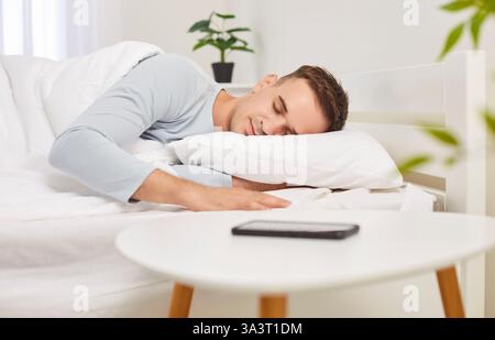 Ritratto di un giovane che dorme sul letto in un'accogliente camera bianca moderna con telefono cellulare sul tavolo. Foto Stock