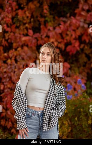 Autunno Ritratto alla moda: Giovane donna con un outfit casual che si posa contro la vivace parete di edera rossa in un ambiente urbano all'aperto. Eleganti vetrine in stile stagionale Foto Stock